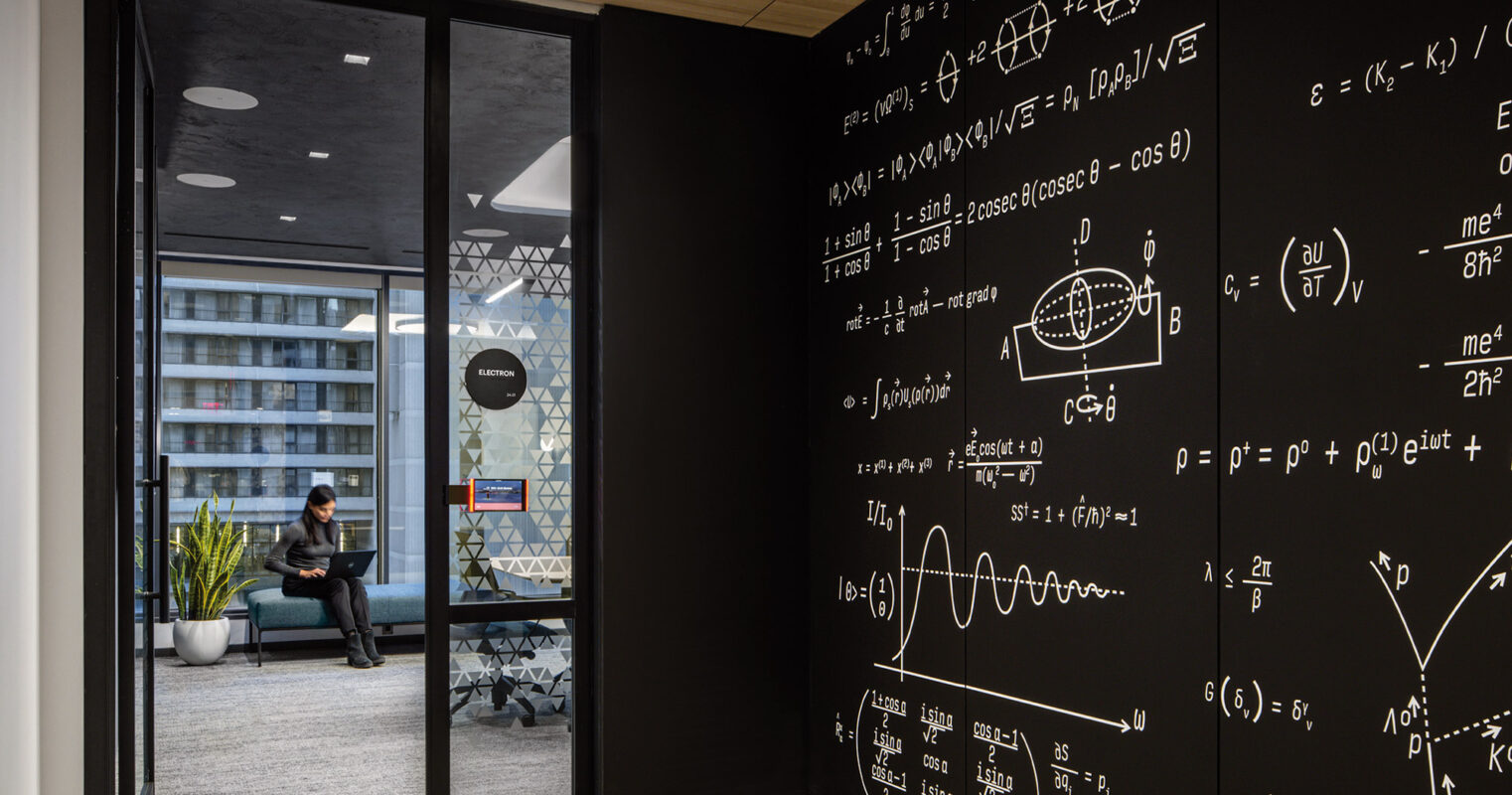 A modern office space with a person sitting in the background. The focal point of the room is a large black wall adorned with intricate white mathematical equations and diagrams.