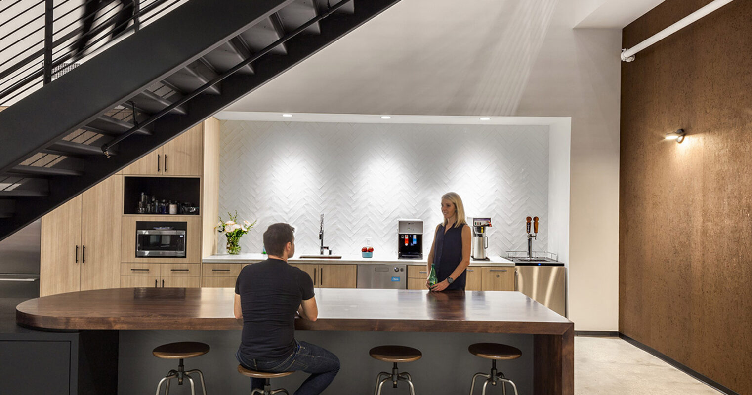 Modern kitchen with an L-shaped wooden island, geometric patterned backsplash, and integrated appliances. High ceilings and a floating staircase complement the space's minimalist aesthetic.