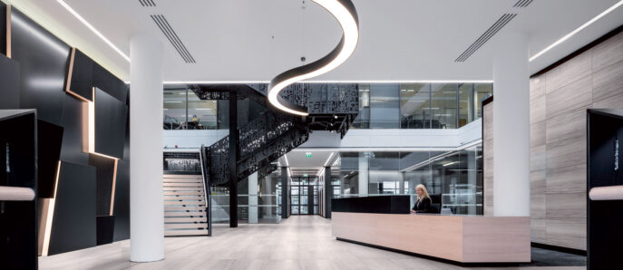 Modern office lobby with a minimalist design featuring sleek, curved lighting overhead, contrasting textures with stone accent walls, wood panel flooring, and a central reception desk with clean lines. A spiral staircase adds a focal point, enhancing the space's vertical dynamics.