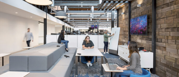 Modern office space with open-floor design, featuring exposed wooden beams and industrial-style lighting. Streamlined white workstations complement the natural wood accent wall, while vibrant artwork adds a pop of color. Comfortable seating spots encourage collaborative and flexible work arrangements.