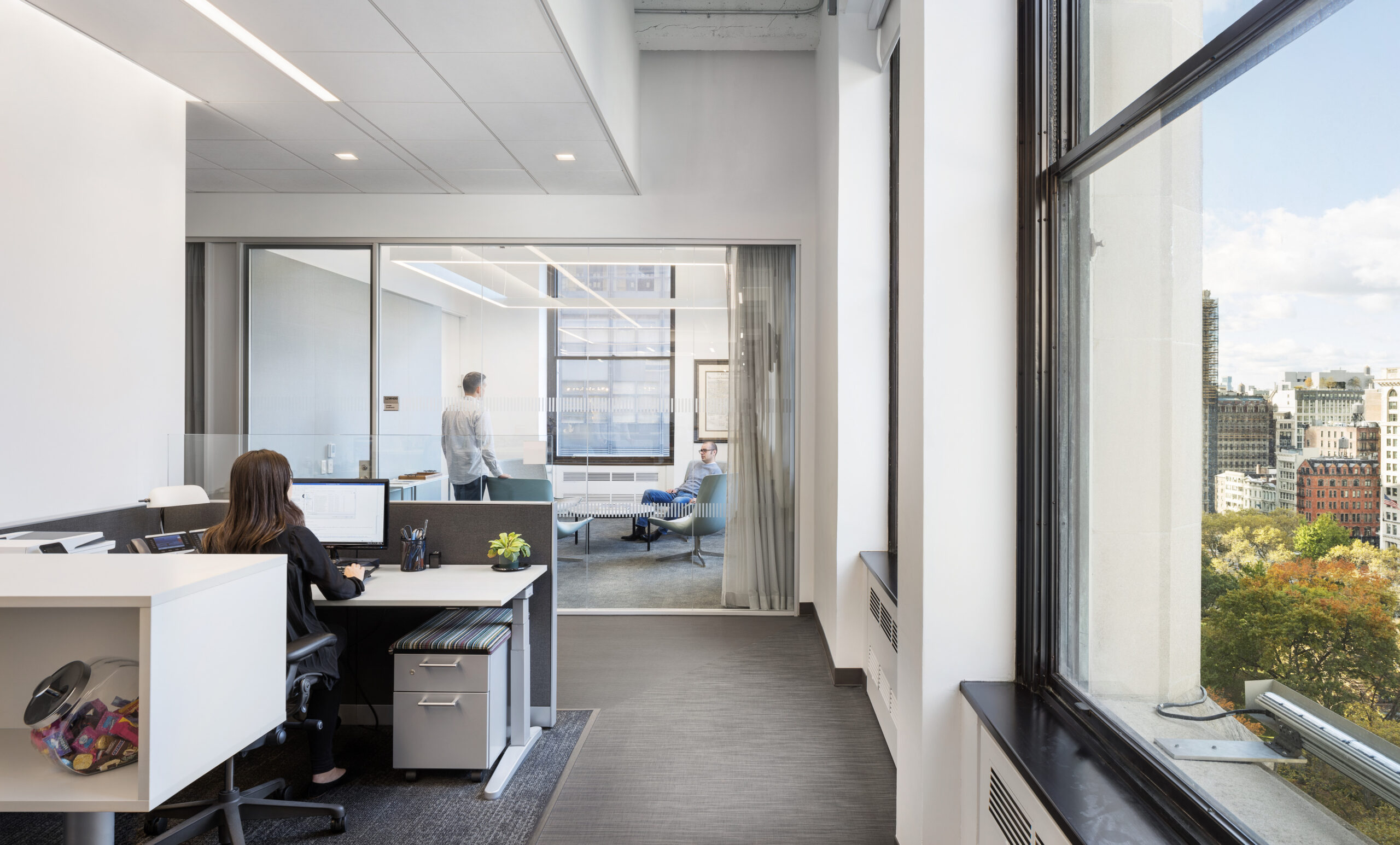 A modern office space featuring a sleek design, with a focused worker at a minimalist desk. Glass meeting rooms are visible, filled with natural light from expansive windows. Neutral colors enhance the sophisticated ambiance, while the city skyline peeks through, adding an urban touch to the professional setting.