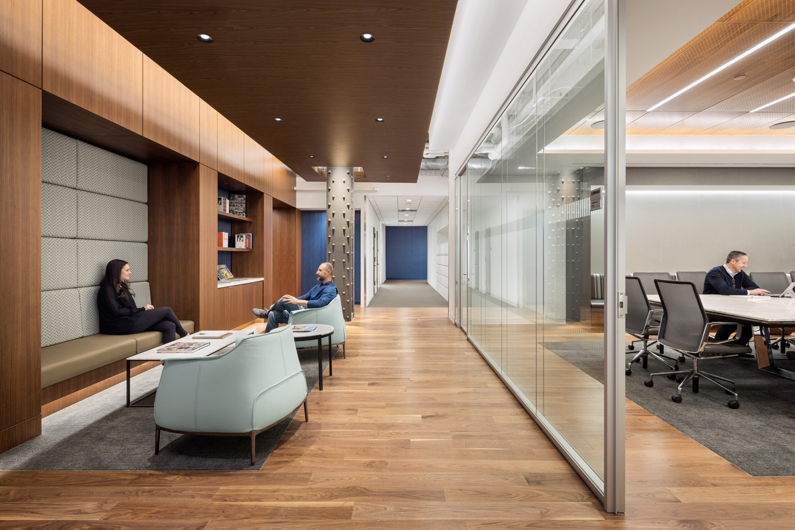 A modern office interior featuring a cozy seating area with a wooden panel backdrop, complemented by frosted glass partitions. Natural wood flooring extends through the space, while contemporary furnishings in soft hues enhance the inviting atmosphere. Employees are engaged in conversation and work, illustrating a collaborative environment.