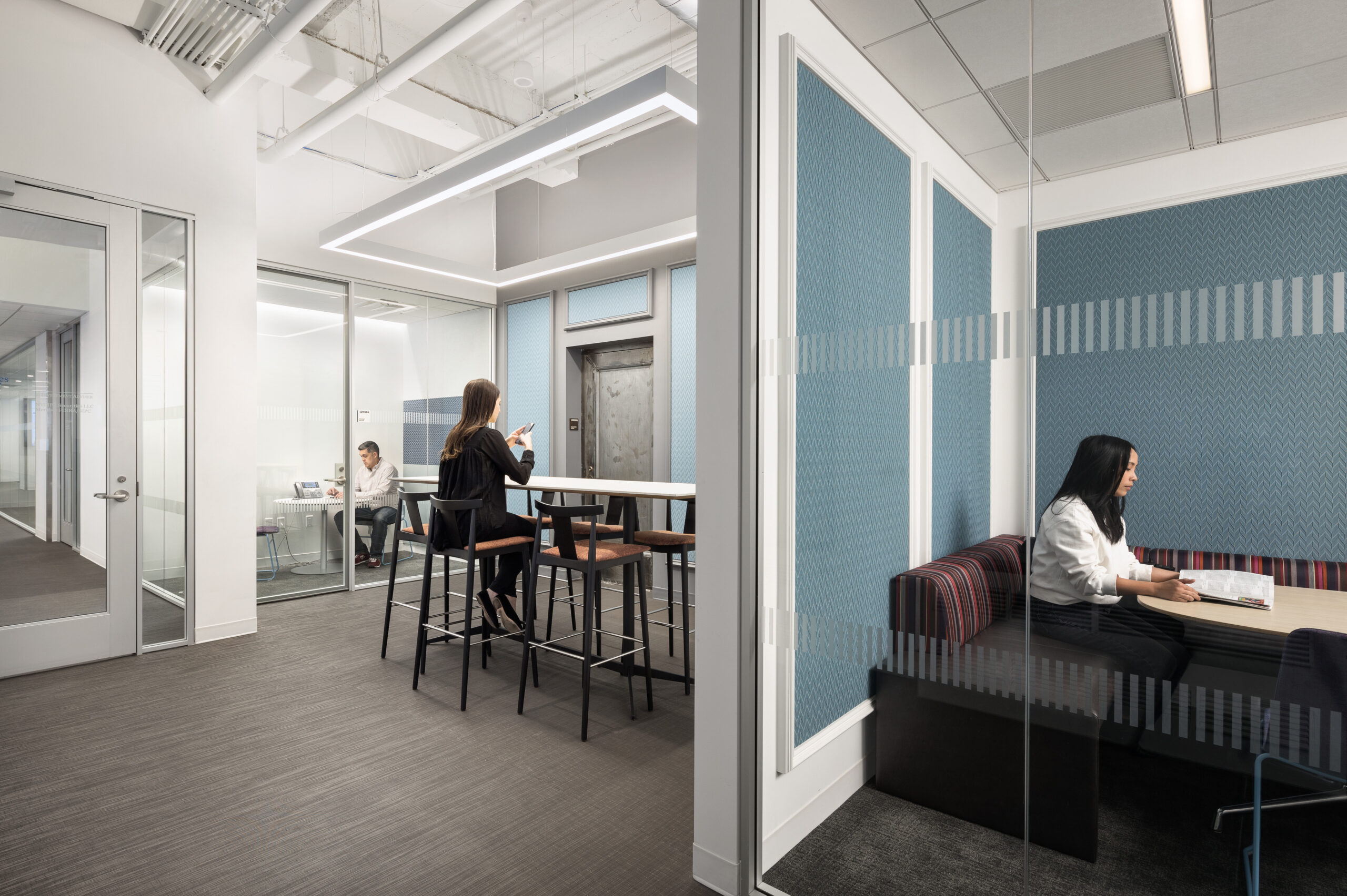 Modern office design highlights open and collaborative spaces. A sleek break area features high stools around a bar-height table, while glass-walled meeting rooms ensure transparency. Subtle blue paneling adds warmth, complemented by textured seating, fostering an inviting atmosphere for creativity and teamwork. Natural light enhances the space’s productivity.