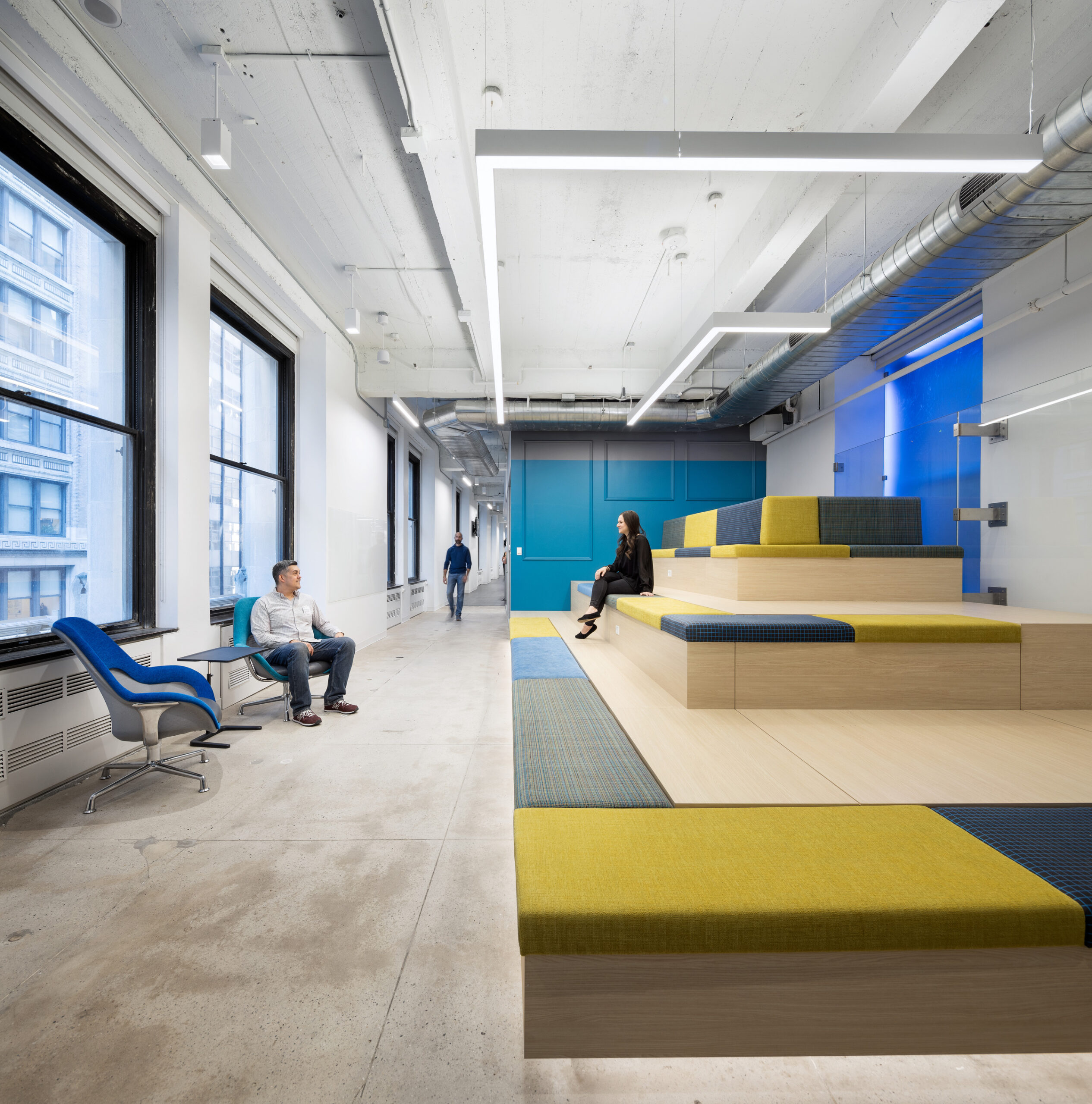 Modern office space featuring tiered seating with vibrant yellow and blue cushions, promoting collaborative work. Large windows allow natural light, enhancing the open layout. Industrial design elements, including exposed concrete and ductwork, blend with contemporary furniture, creating a dynamic and functional environment for creativity and interaction.