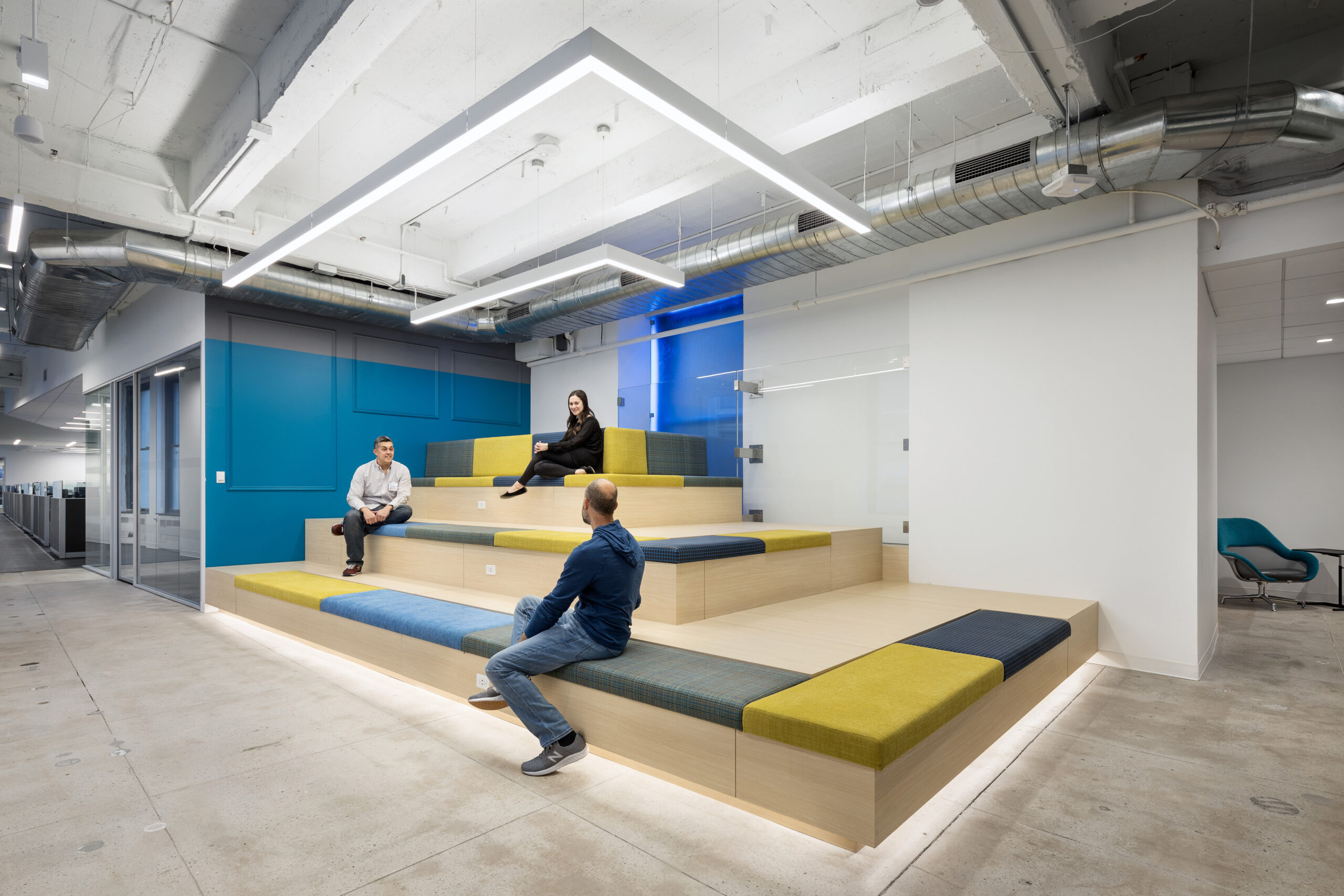 A modern collaborative space featuring tiered wooden seating with vibrant green and blue cushions. Individuals engage in conversation while illuminated by sleek, linear LED lighting above. The industrial ceiling with exposed ductwork enhances the contemporary atmosphere, promoting creativity and teamwork in this thoughtfully designed environment.