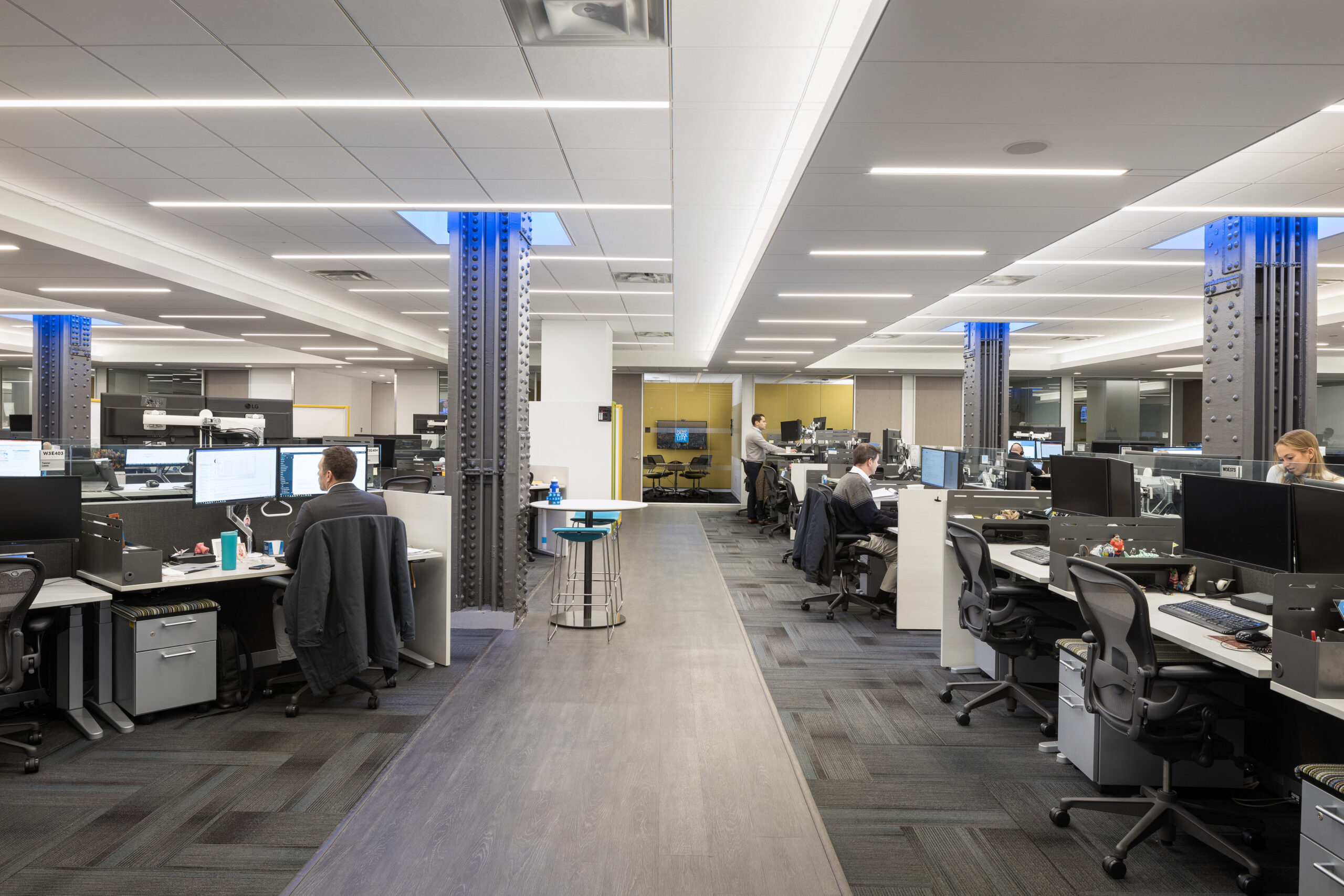 Modern office layout featuring open workstations arranged in collaborative clusters, defined by sleek lines and ergonomic furniture. Industrial-style metal columns complement the contemporary design, while soft lighting enhances productivity. A neutral color palette with touches of blue creates a professional yet inviting atmosphere, promoting comfort and efficiency for employees.