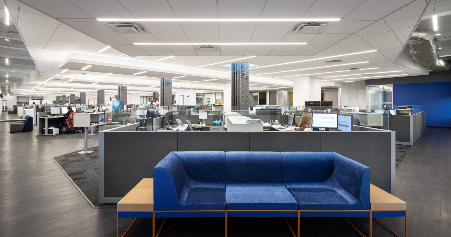 Modern open office layout showcasing ergonomic workstations surrounded by sleek cubicles. A vibrant blue sofa with wooden accents serves as a collaborative seating area. Soft LED lighting enhances the spacious design, promoting productivity and comfort while allowing for seamless communication among team members.
