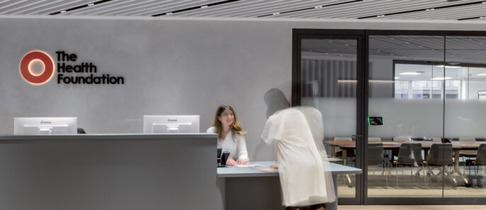 Modern health foundation office lobby with sleek, horizontal lines emphasizing the linear design. Wooden slats adorn the ceiling, while the reception desk's minimalist gray tone contrasts with the warm lighting. Staff interacts professionally, providing a welcoming atmosphere amidst a backdrop of transparent meeting rooms.