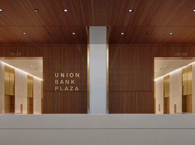 Modern and minimalist reception area featuring a sleek white desk against a textured wooden slat backdrop. Overhead linear lighting accents the warm tones, and recessed floor lighting adds a dramatic effect near the entrance marked "Union Bank Plaza".