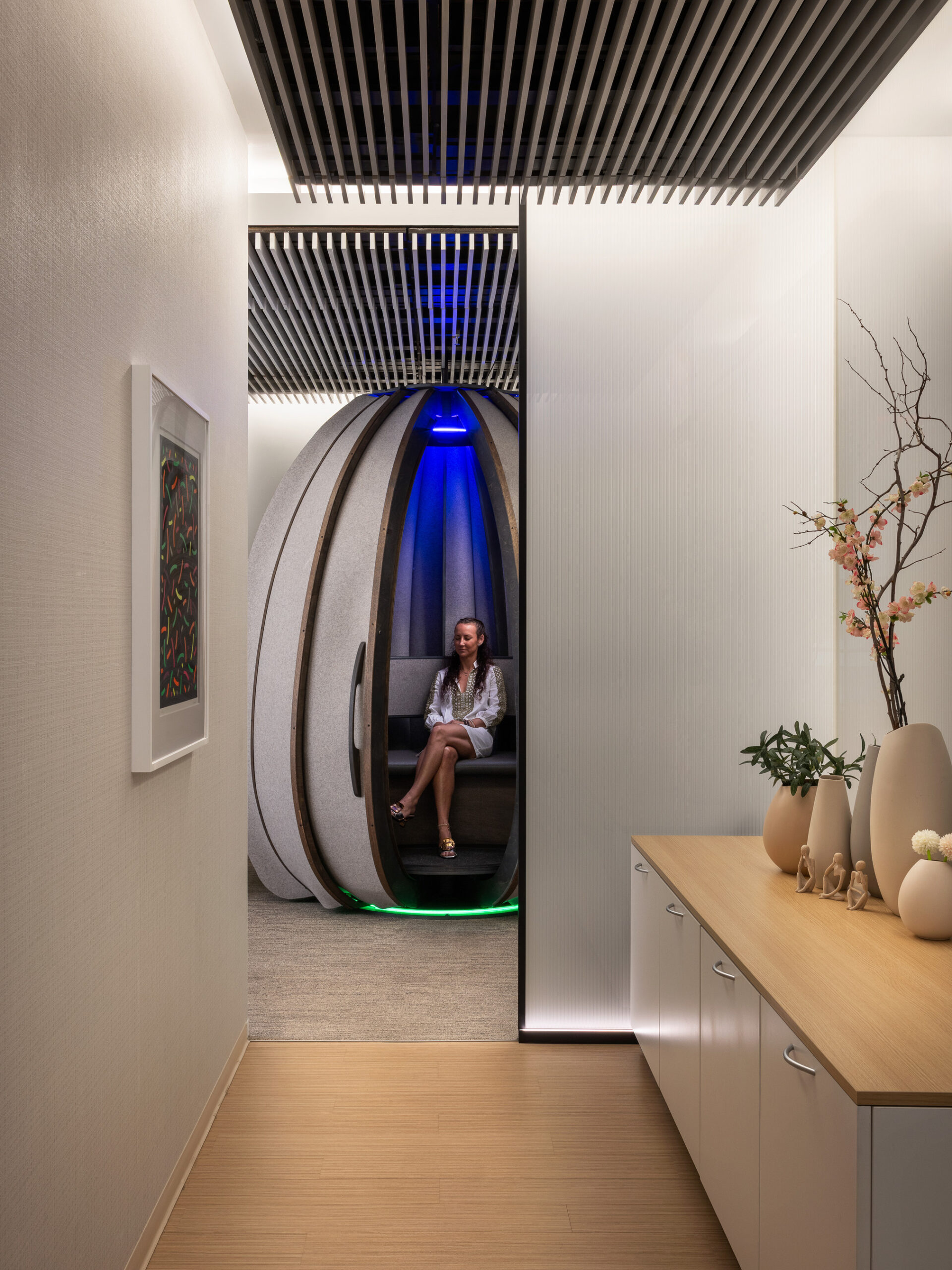 A modern interior space features a sleek hallway leading to a unique pod-like structure with soft, ambient lighting. A woman relaxes inside the pod, demonstrating innovative use of minimal design elements. Natural materials and a light color palette enhance the serene atmosphere, promoting both comfort and creativity.