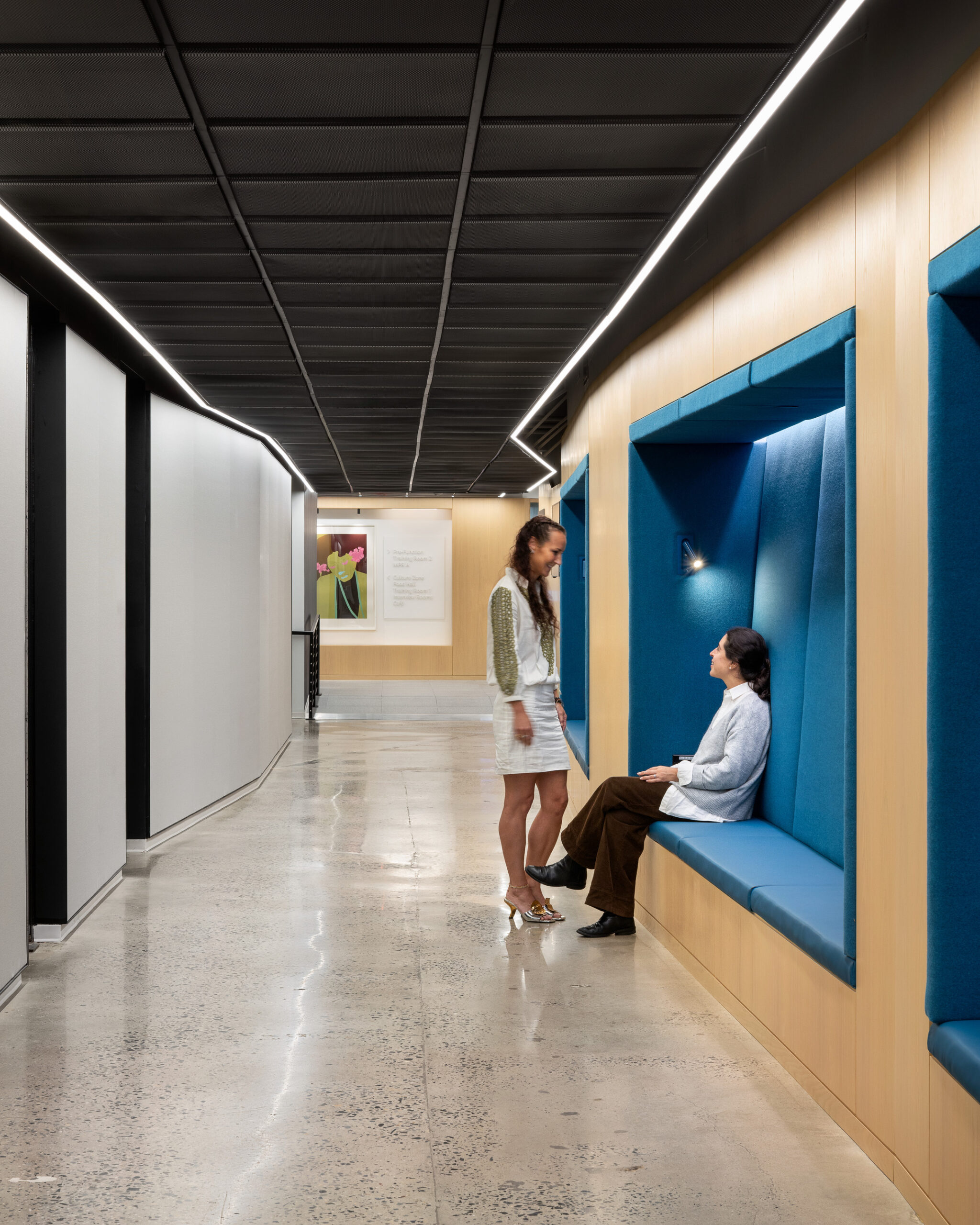 A contemporary corridor features sleek, polished concrete flooring and textured wooden paneling. On the left, a woman engages in conversation with a seated individual in a modern blue upholstered recess. Soft, ambient lighting from overhead fixtures enhances the inviting atmosphere, promoting collaboration and comfort within the space.