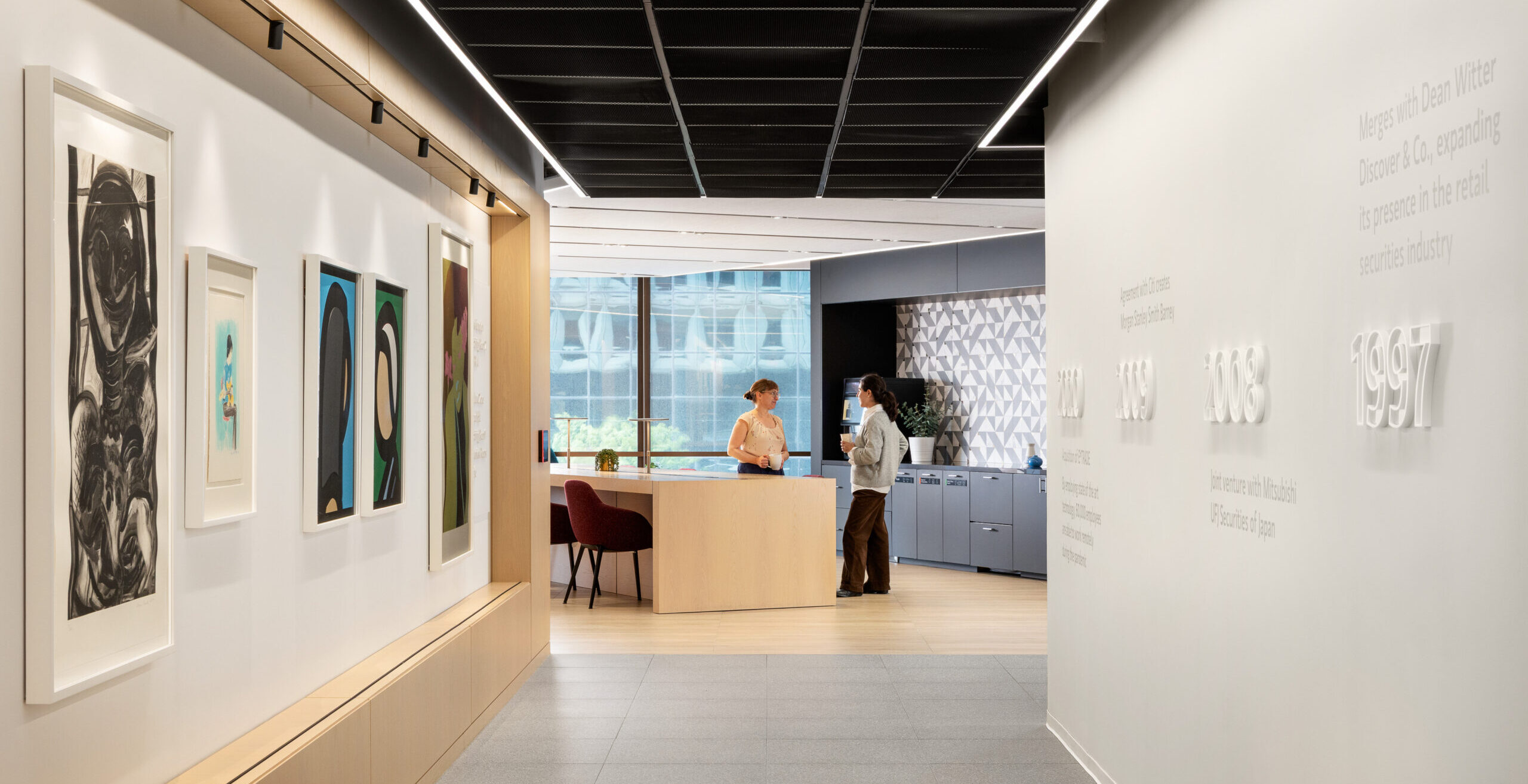 Brightly lit hallway featuring a modern reception area set against a backdrop of abstract artwork. Sleek wall design combines wood paneling with a minimalist aesthetic. Individuals engage in conversation, embodying a welcoming atmosphere that emphasizes collaboration and contemporary design in a professional setting. Textural contrasts enhance visual interest.