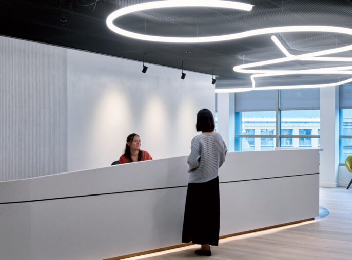 Modern office lobby with circular overhead LED lighting, streamlined reception desk, wooden flooring, and pops of color from yellow carpet and blue-cushioned chairs in the seating area.