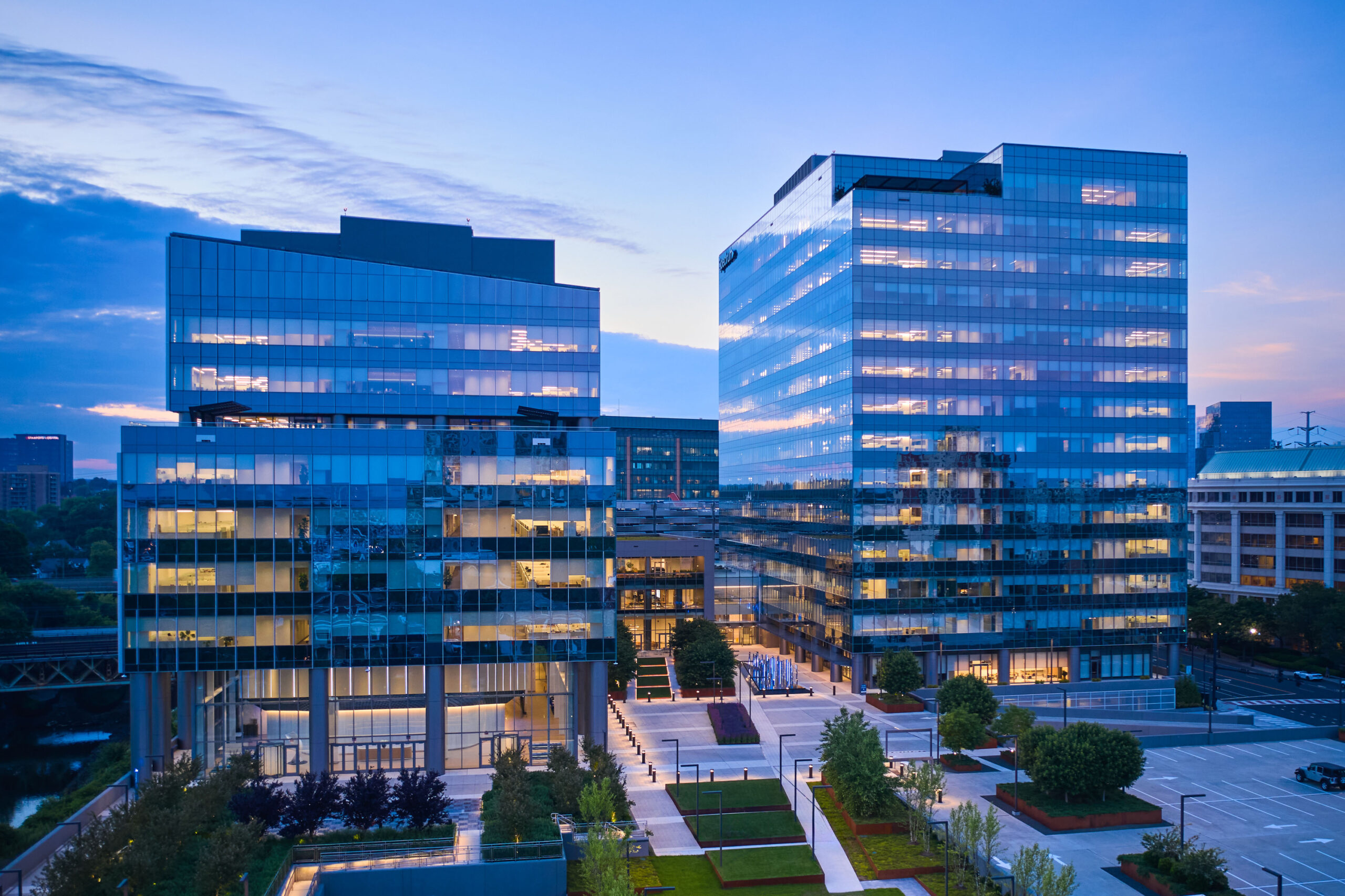 Two modern glass-clad skyscrapers reflect the vibrant colors of the sunset. The design features sleek lines and expansive windows, showcasing a contemporary aesthetic. Surrounding landscaped areas include greenery and seating spaces, enhancing the urban environment while promoting connectivity and collaboration among the buildings.