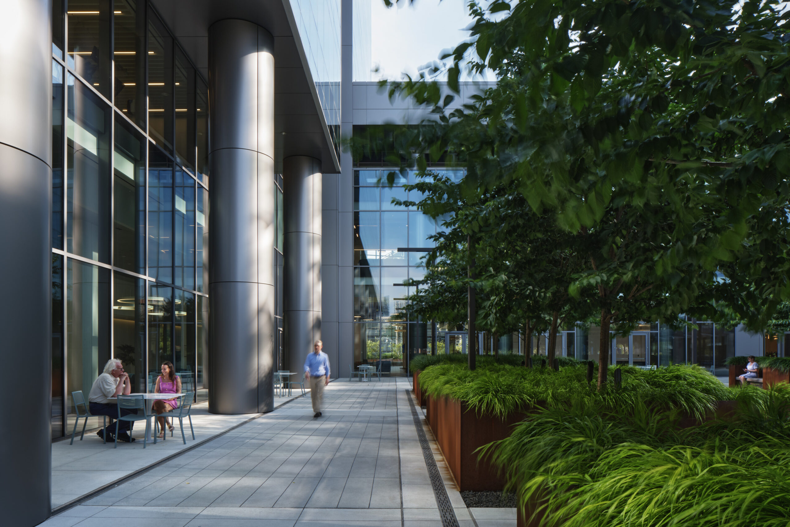 A sleek, contemporary courtyard features large glass panels reflecting natural light, framed by polished metal columns. Lush greenery and low-maintenance plants enhance the space, while seating areas invite casual gatherings. A pathway leads through the setting, creating a harmonious blend of architecture and nature in this urban environment.