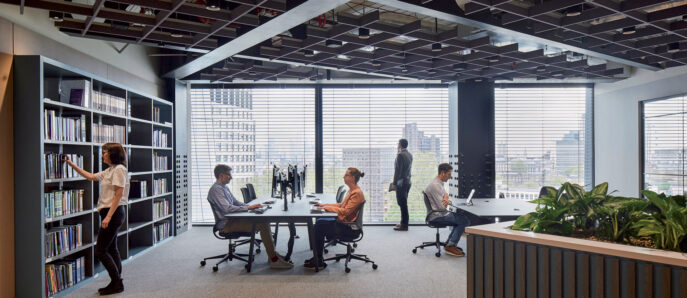 Modern open-plan office space with floor-to-ceiling windows offering cityscape views. Features include a large bookshelf, collaborative workstations, a green wall divider, and exposed ceiling beams, fostering a blend of industrial and biophilic design elements.