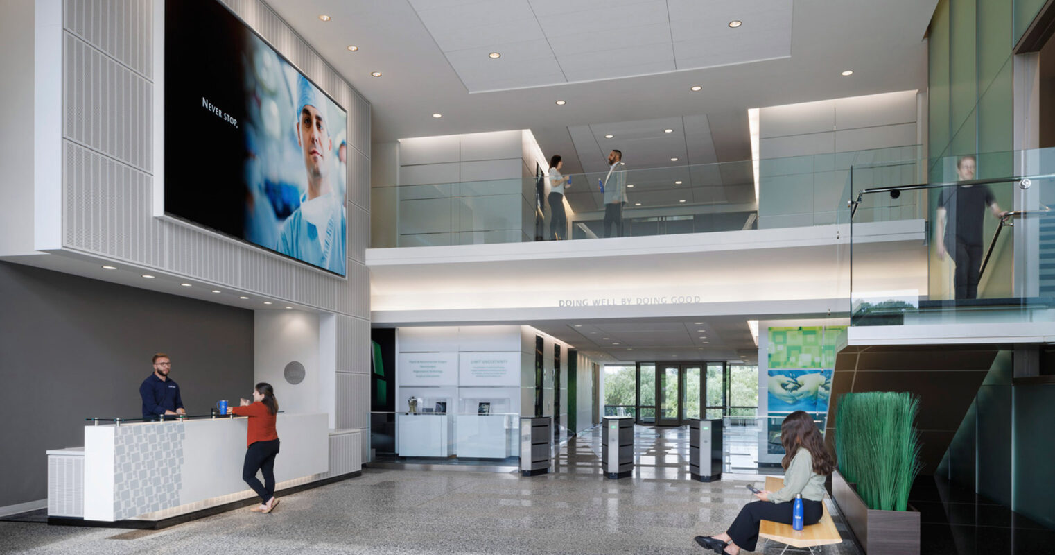 Spacious corporate lobby featuring a minimalist reception desk, large digital display screen, and glass-paneled second-story walkway. The design exudes a modern aesthetic with clean lines, ample natural light, and a monochromatic color scheme complemented by greenery and contemporary seating areas.