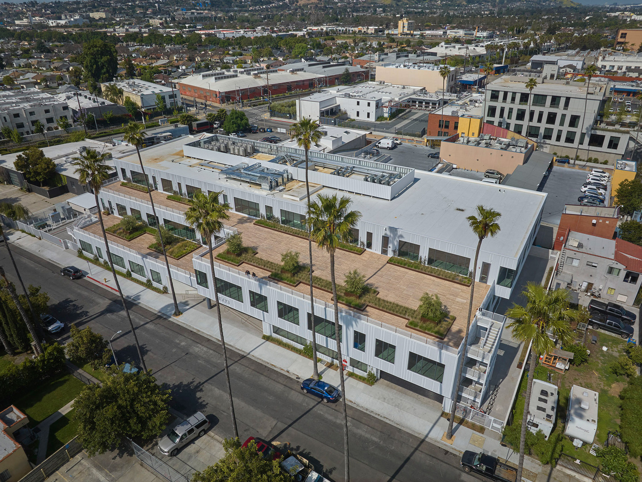 Bird's eye view of a modern, multi-story residential building featuring clean lines, flat rooftops, and lush greenery on balconies; nestled in a mixed urban landscape. The contemporary architecture presents a harmonious blend with the surrounding commercial structures.