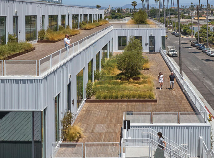 Modern commercial architecture featuring a modular building design with white corrugated metal exteriors. Landscaped green roof terraces and external staircases complement the urban backdrop, enhancing sustainability and communal space.