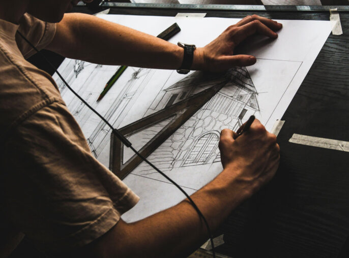 Overhead view of an architect's hands drawing a detailed architectural sketch of a classic house with a gabled roof and arched windows, using a straightedge and pencil on paper, conveying a sense of design precision and creativity.