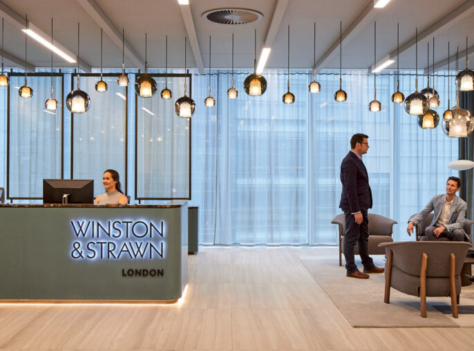 Modern corporate lobby with light wood floors, a sleek reception desk, suspended glass pendant lights, and seated area with neutral-toned furniture. Large windows with sheer curtains provide natural light, complemented by recessed and accent ceiling lighting.