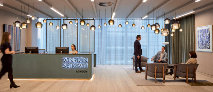 Modern corporate lobby with light wood floors, a sleek reception desk, suspended glass pendant lights, and seated area with neutral-toned furniture. Large windows with sheer curtains provide natural light, complemented by recessed and accent ceiling lighting.