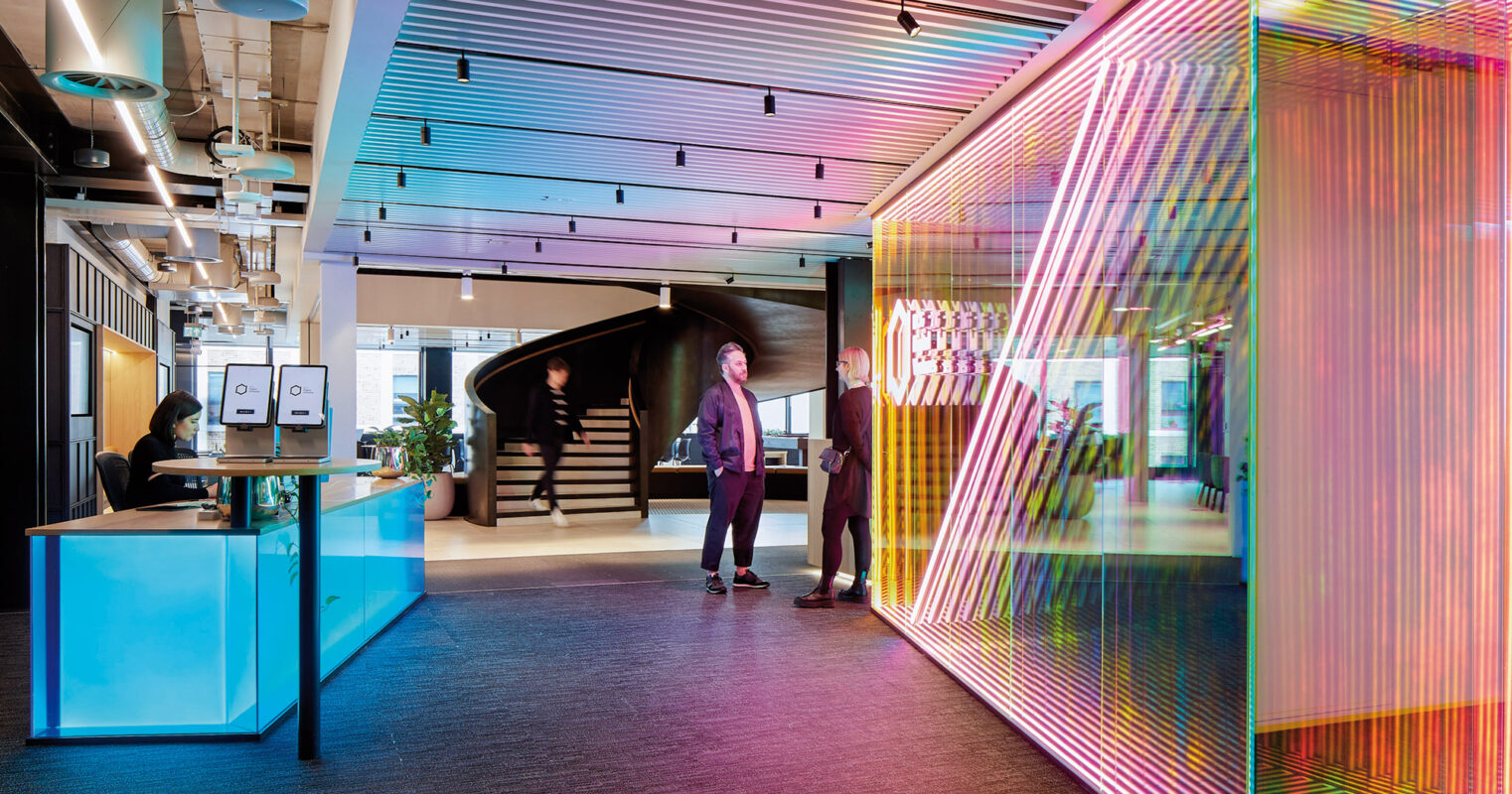 A modern office space featuring a vibrant, colorful glass-walled meeting room, with individuals engaging in work activities in a contemporary, well-lit interior.