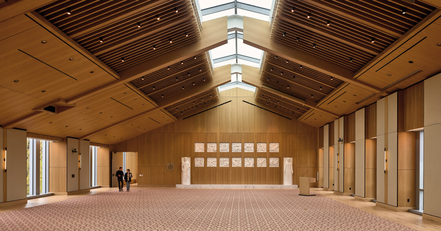 Elegant modern interior of a spacious hall with wooden paneled ceiling, patterned wall decorations, and geometric carpet design, with two people conversing near the entrance.