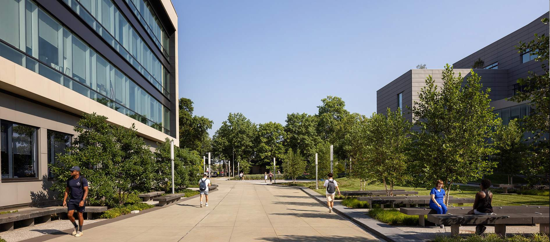 Pedestrians walking along a pathway flanked by modern buildings and green landscaping on a sunny day.