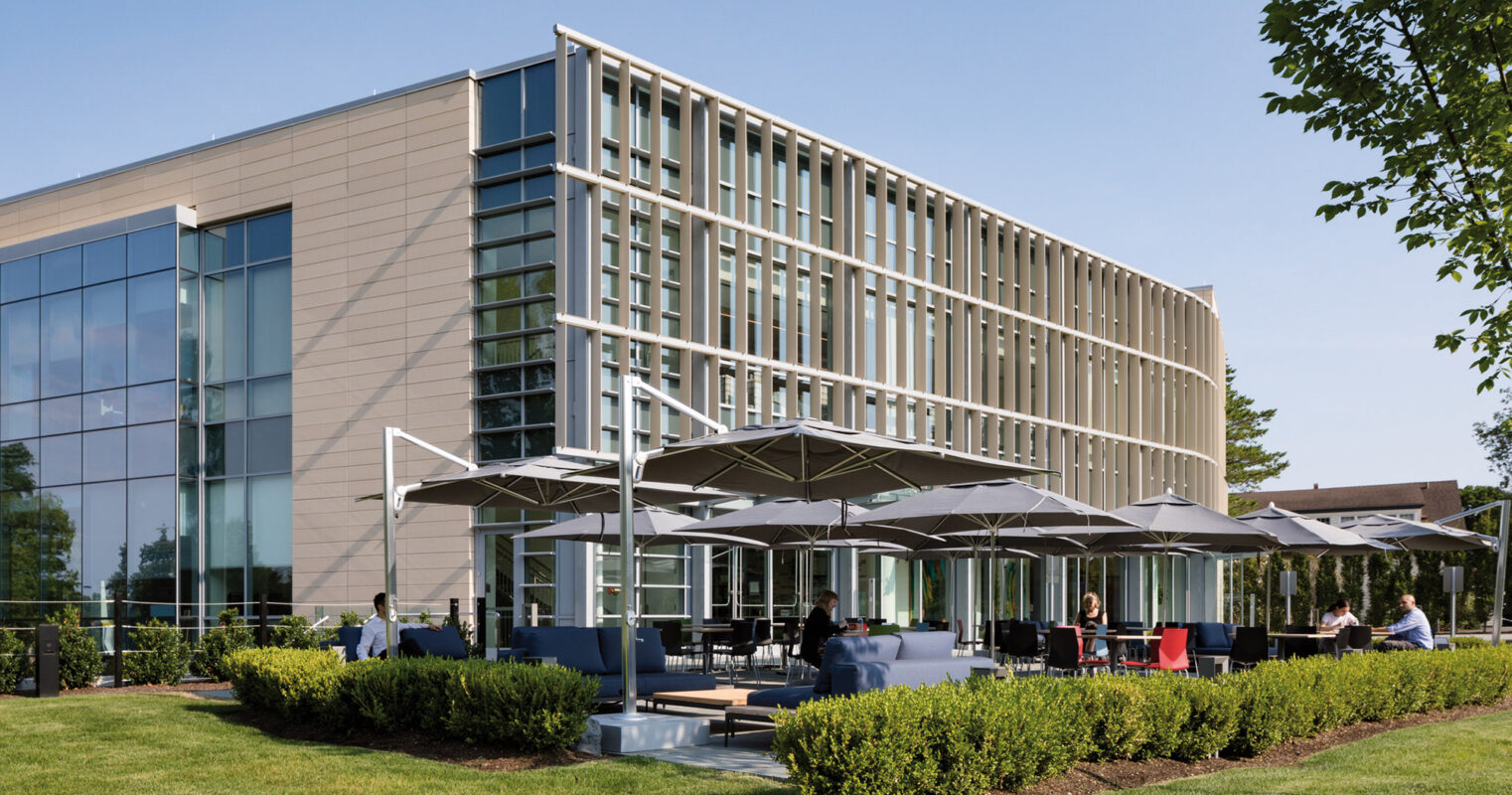 Modern office building with an outdoor seating area featuring umbrellas and tables, set amidst manicured lawns on a sunny day.