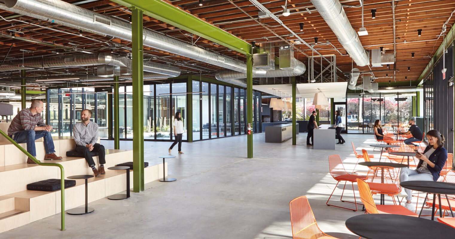 Modern office break area with industrial-style exposed ceiling, vibrant orange chairs, and employees enjoying a casual conversation.