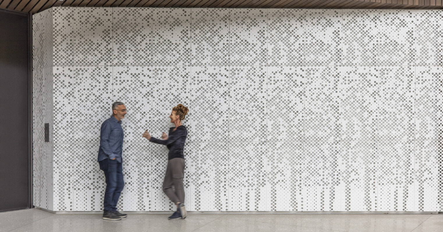 Two individuals converse in a modern hallway featuring a geometric-patterned perforated metal screen wall, providing both privacy and light diffusion, accented by a warm wood ceiling and minimalist design.