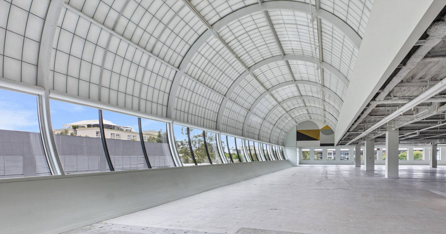 Spacious modern interior with arched ceiling, featuring expansive translucent panels providing abundant natural light, and a mixture of concrete finishes with geometric color accents, emphasizing sleek architectural lines.