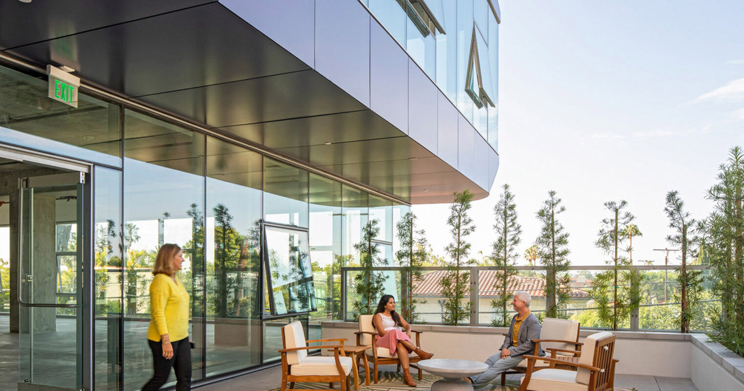 Modern outdoor terrace with wooden furnishings, glass-paneled railings, and a lush green backdrop. The setting features an informal meeting area with minimalist design, emphasizing open space and integration with nature.
