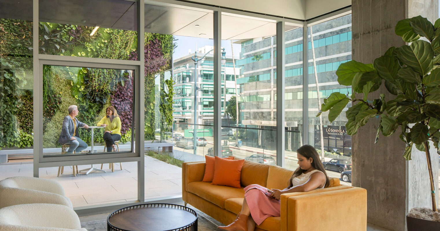 Spacious lobby with floor-to-ceiling windows, allowing natural light to highlight the earth-toned velvet sofa and lush indoor greenery. Concrete walls complement the clean, modern aesthetic, with a view of the urban environment blending seamlessly with the interior space.