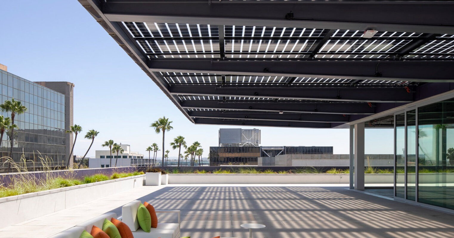 Spacious outdoor terrace with sleek, white modular seating under a modern black pergola. The overhead slats cast geometric shadows, juxtaposing the bright midday sun, with a skyline view providing an urban backdrop. Vibrant green planters add a touch of nature to the minimalist design.