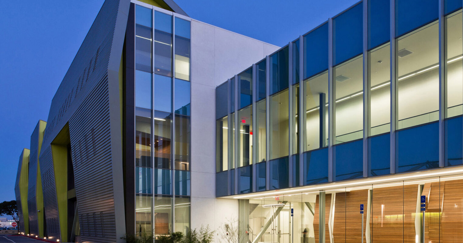 Modern architectural building at twilight featuring angular design and asymmetrical windows. The facade combines reflective glass with warm wooden panels above a concrete base, and includes a glass entrance canopy.