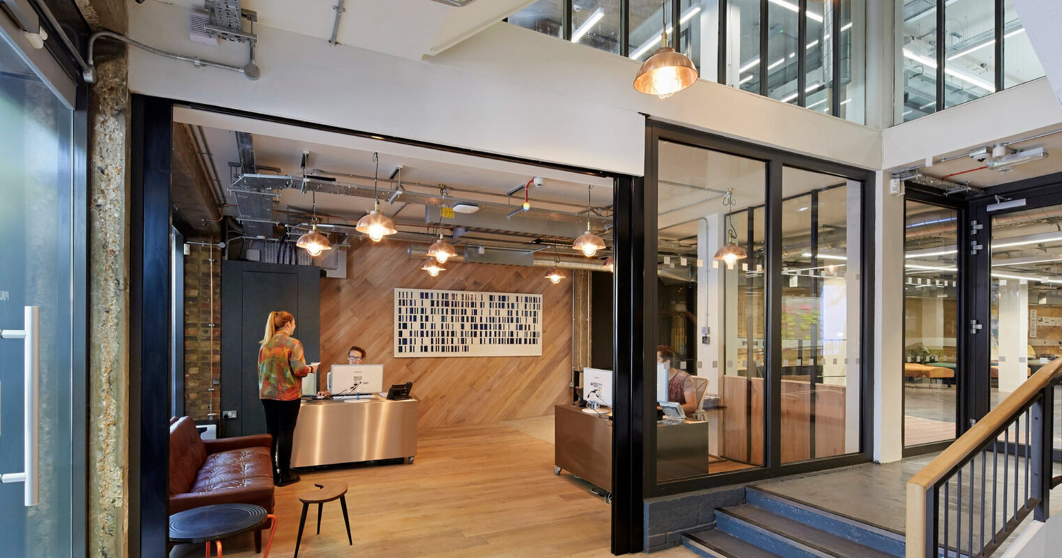 Modern open-plan office space blending industrial and contemporary styles, featuring exposed ductwork, concrete flooring, and pendant lighting. Glass partitions provide transparency while maintaining distinct work areas, complemented by wooden accents and a welcoming reception desk.