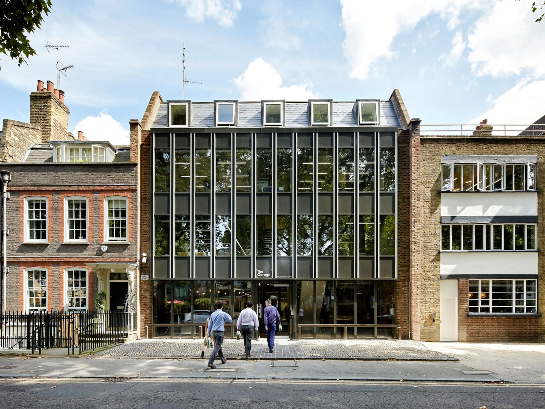 Front view of a contemporary building with a glass facade and geometric window frames nestled between traditional brick structures, integrating modern design with historical architecture.