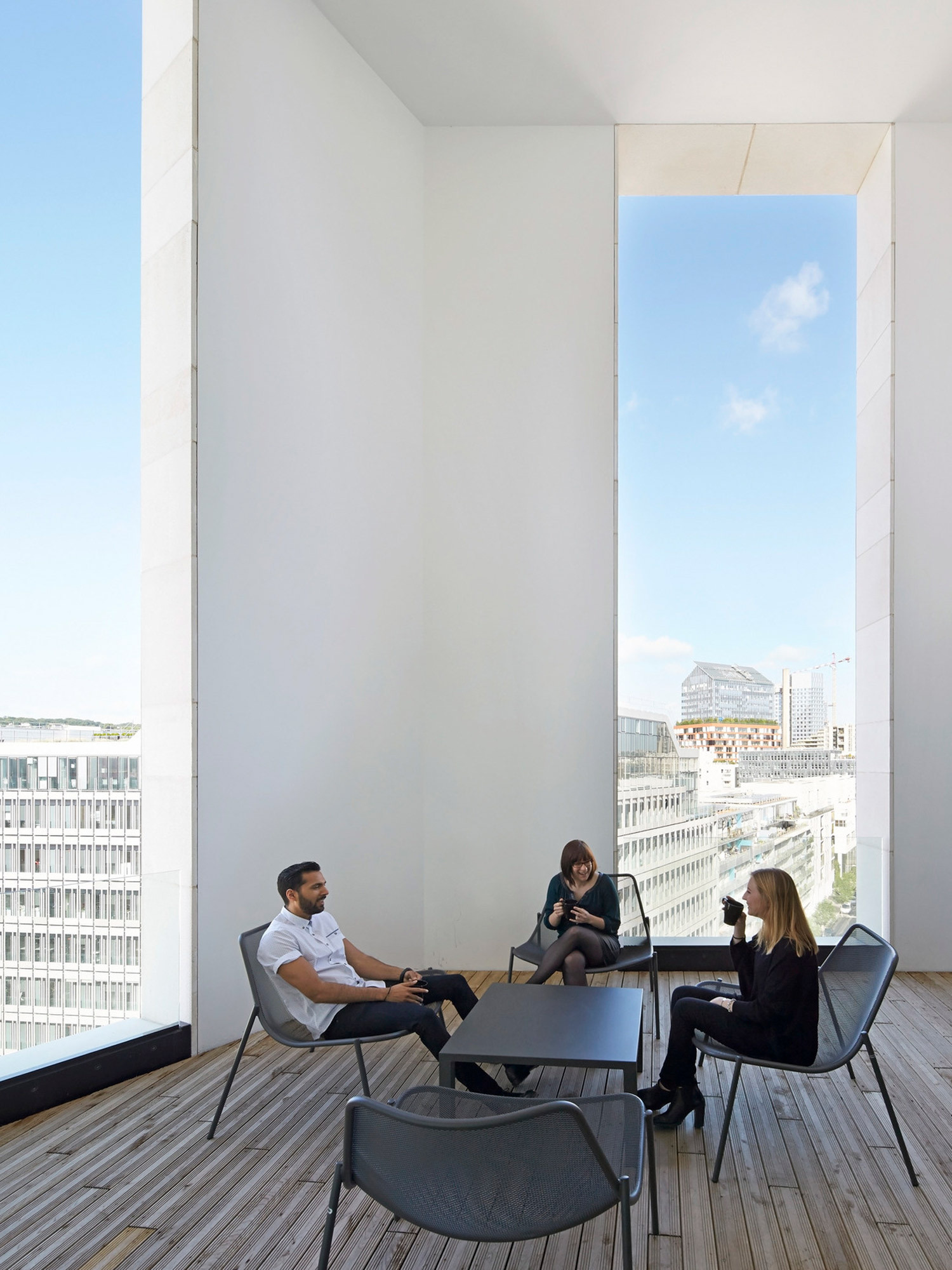 Minimalist interior with full-height glazing offering city views, featuring wood flooring, white walls, and a simple group of modern chairs fostering an informal meeting space.
