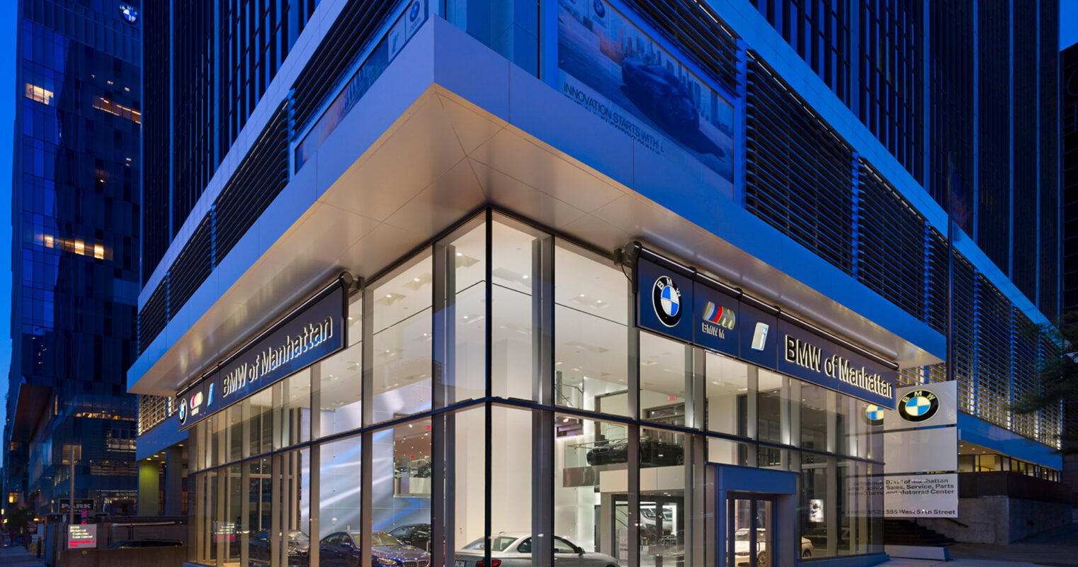 Modern BMW car dealership exterior at twilight with illuminated facade showcasing luxury vehicles through expansive glass windows, flanked by sleek, dark vertical cladding and set against an urban architectural backdrop.