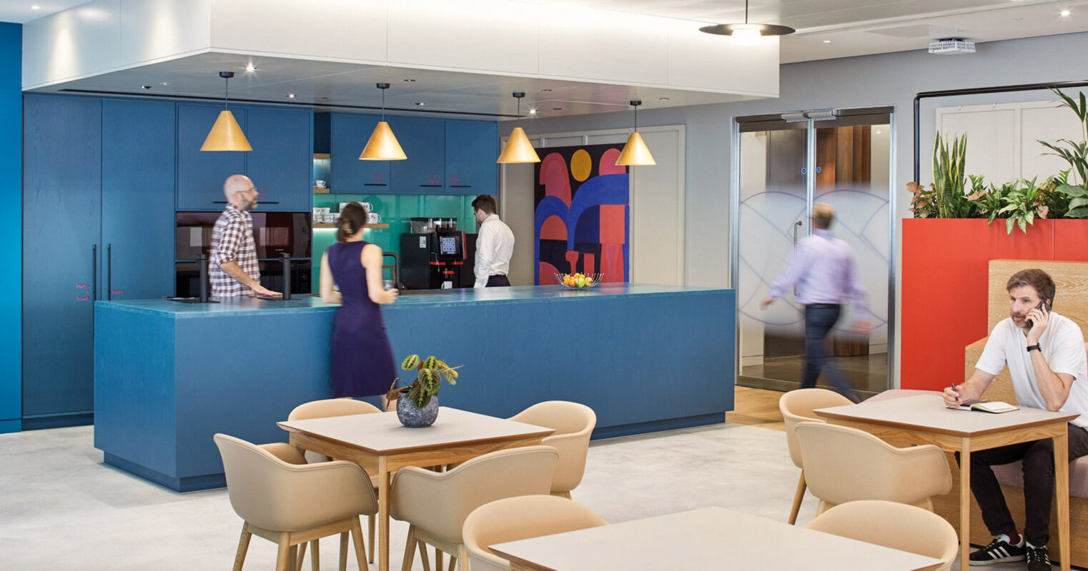 Modern office kitchenette and casual meeting area with light wood tables, white chairs, and pendant lighting. A bold blue color scheme is accented by vibrant wall art, showcasing a clean and contemporary design ethos.