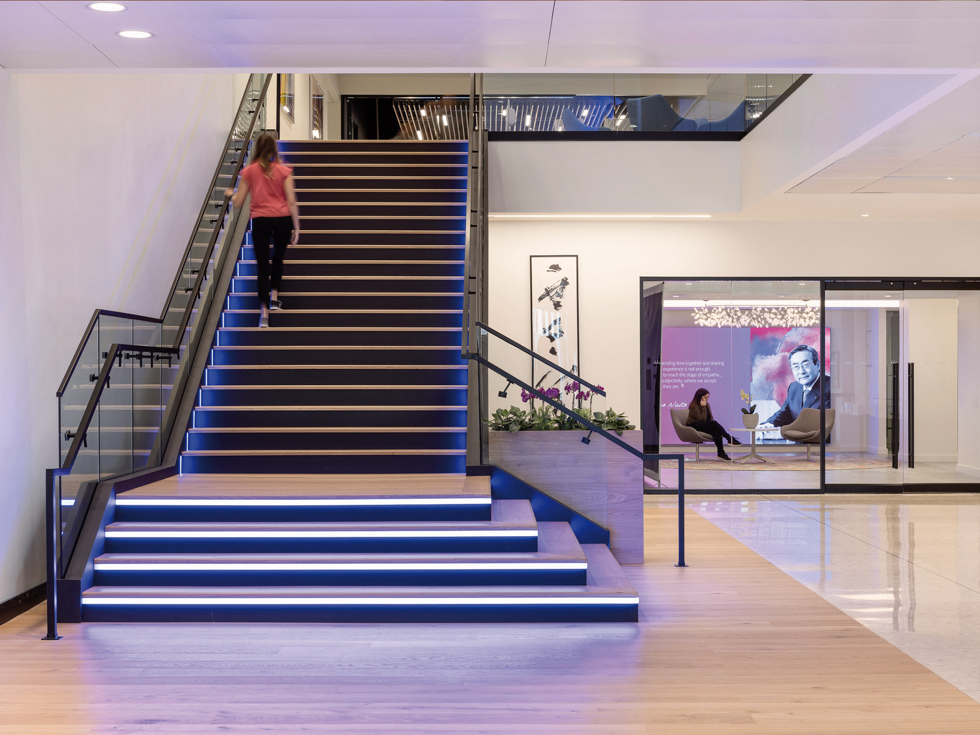 A modern, well-lit interior space featuring an illuminated staircase with a person ascending and another person seated at the bottom, with a large video screen displaying an image of a man in the background.