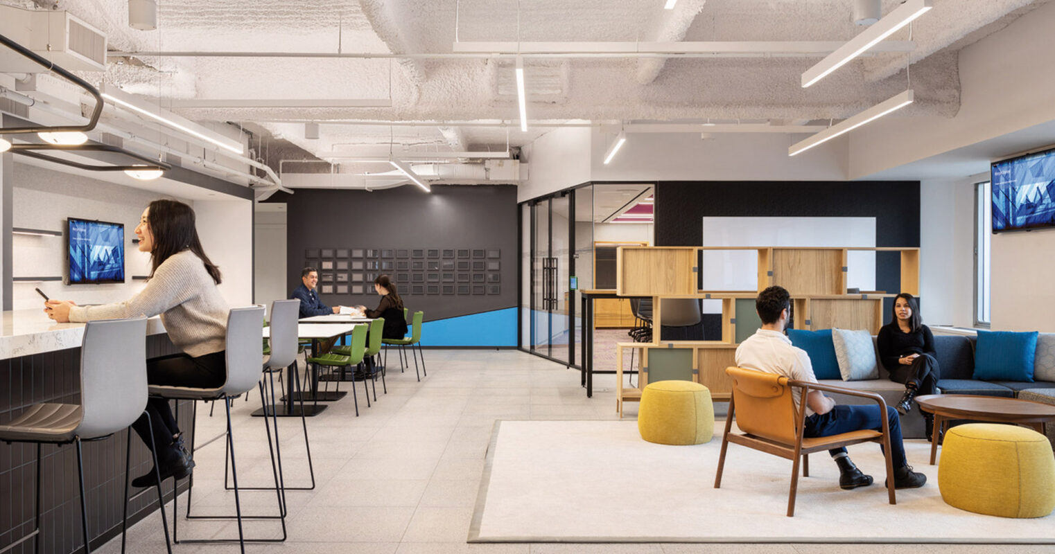 Modern office break area featuring an open-concept layout with exposed white beams, ambient lighting, and a variety of seating styles including high stools and plush circular ottomans. The space also includes wall-mounted monitors and wooden shelving for a functional yet stylish environment.