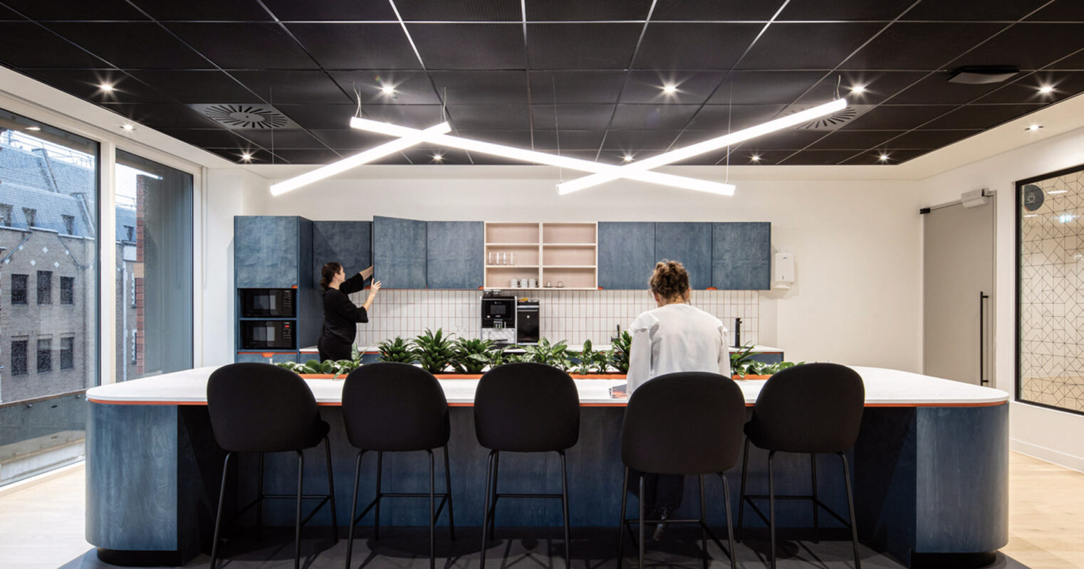 Modern office kitchenette with a sleek, two-toned central island and bar seating, featuring geometric ceiling patterns and ambient lighting. The space combines wood textures and monochrome backdrops to create a welcoming communal area.