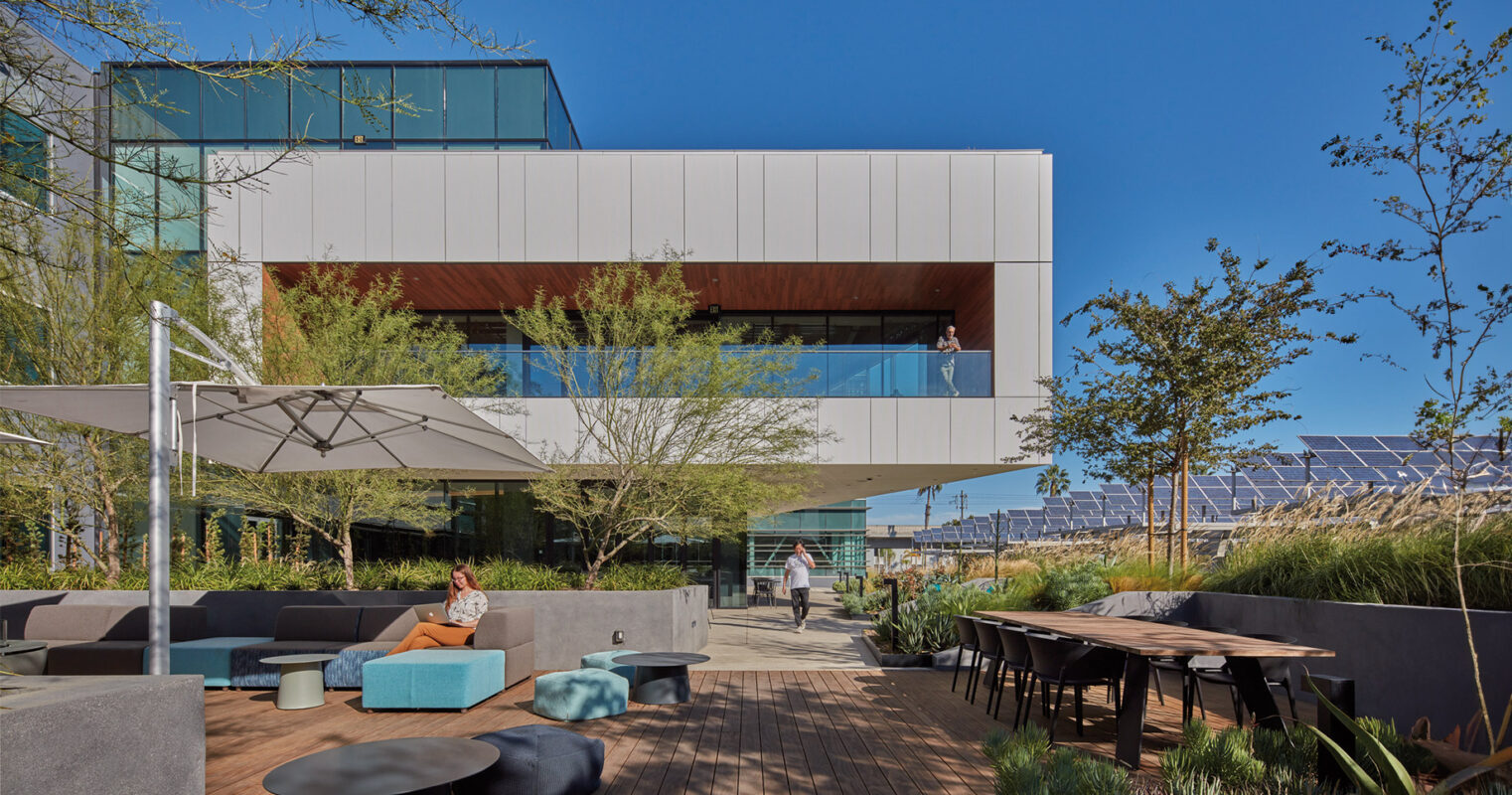 Modern office building with a spacious outdoor seating area featuring a variety of furniture, surrounded by greenery under a clear blue sky.