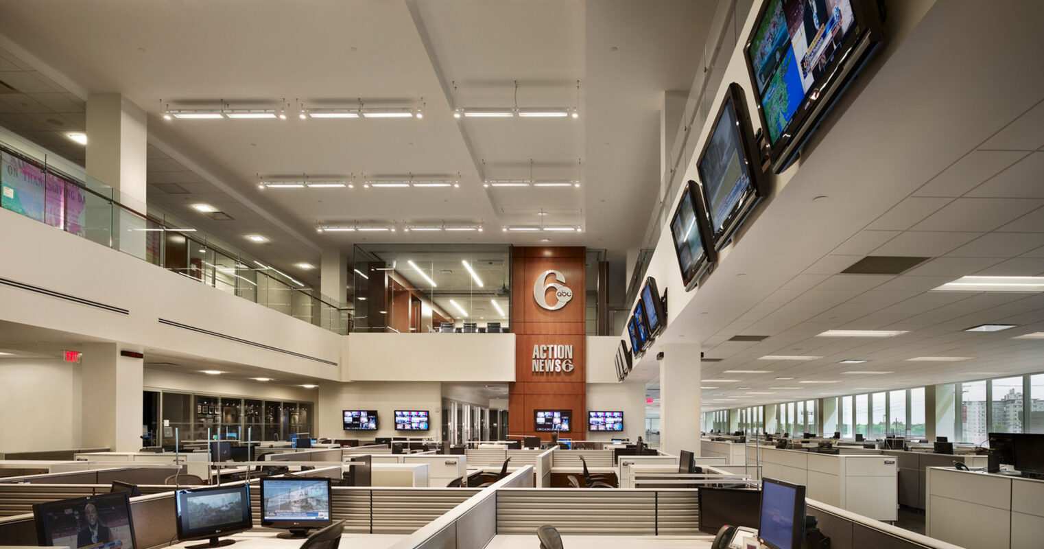 Open-plan office space with rows of workstations under bright, linear pendant lights. Monitors align the central column, complementing the modern, functional aesthetic. A large screen anchors the space above, symbolizing connectivity in a technologically advanced workplace environment.