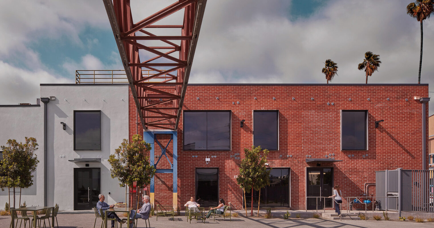 An urban oasis where contemporary architecture meets industrial design, featuring locals enjoying an outdoor seating area under the geometric embrace of an overhead red steel structure, with a serene backdrop of blue skies and palm trees.