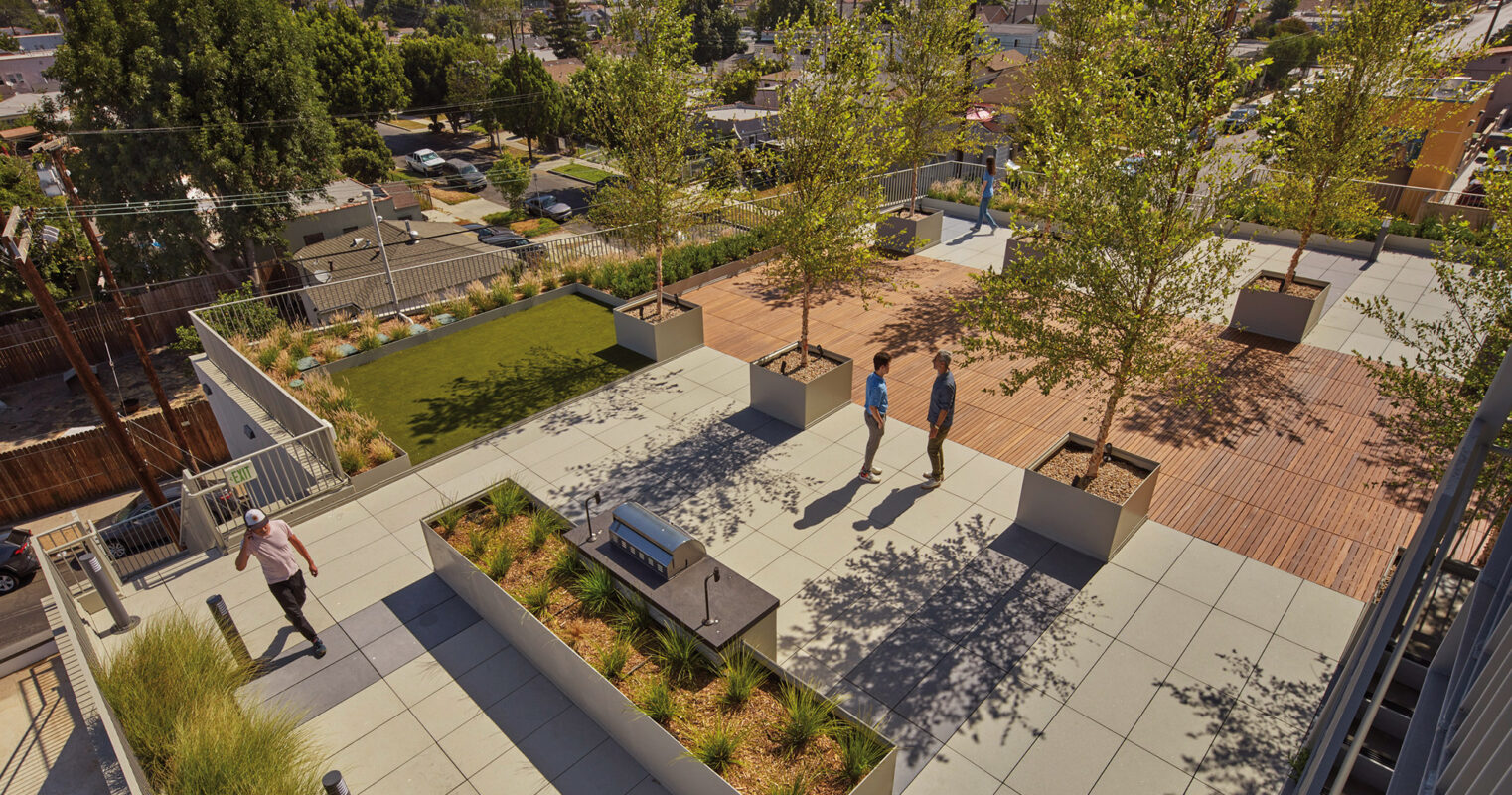 An elevated view of an urban park with geometric landscaping, featuring people enjoying the outdoor space amidst modern benches and lush greenery, with a backdrop of palm trees and a clear blue sky.
