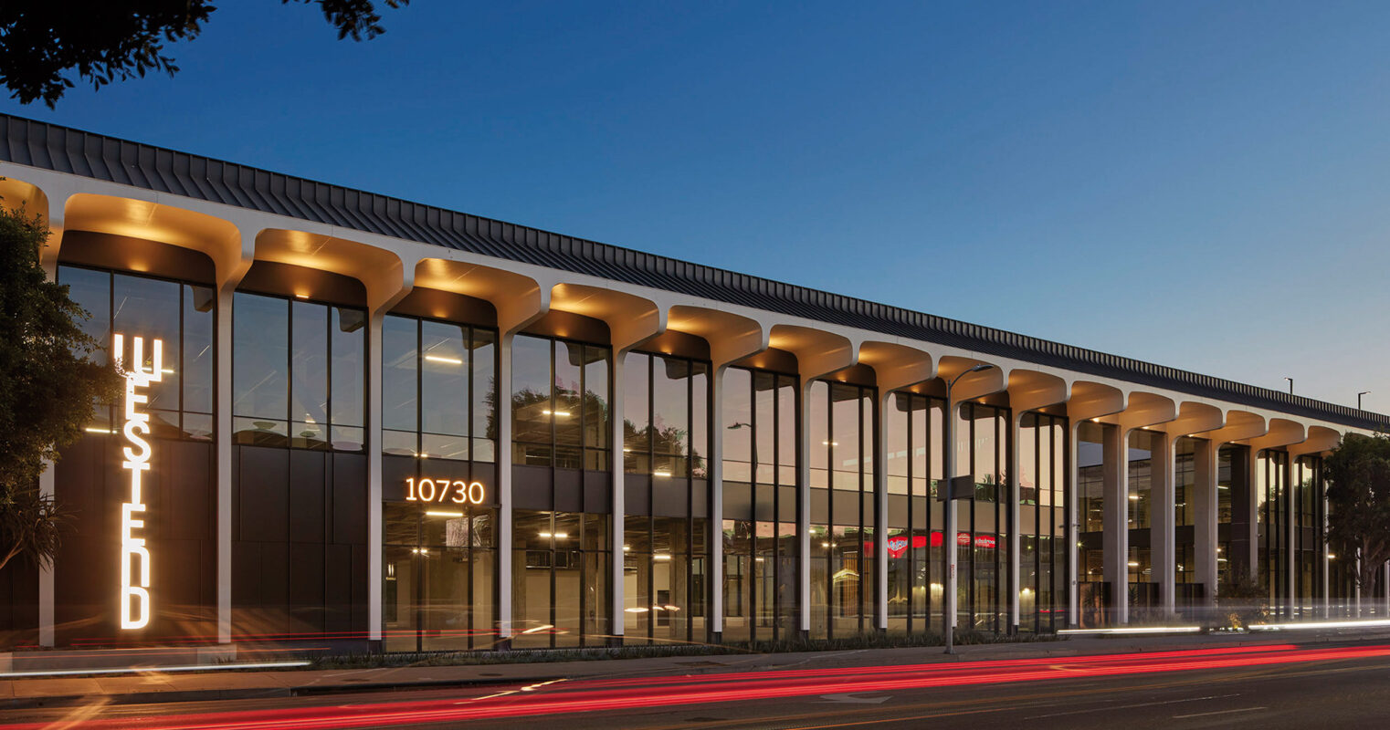 Modern commercial building with elegant columns and large windows illuminated at twilight, with the urban life's streaks of light from passing cars on the bustling street.